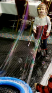 A toddler watches a bubble show, looking a performer make a bubble that is larger than she is